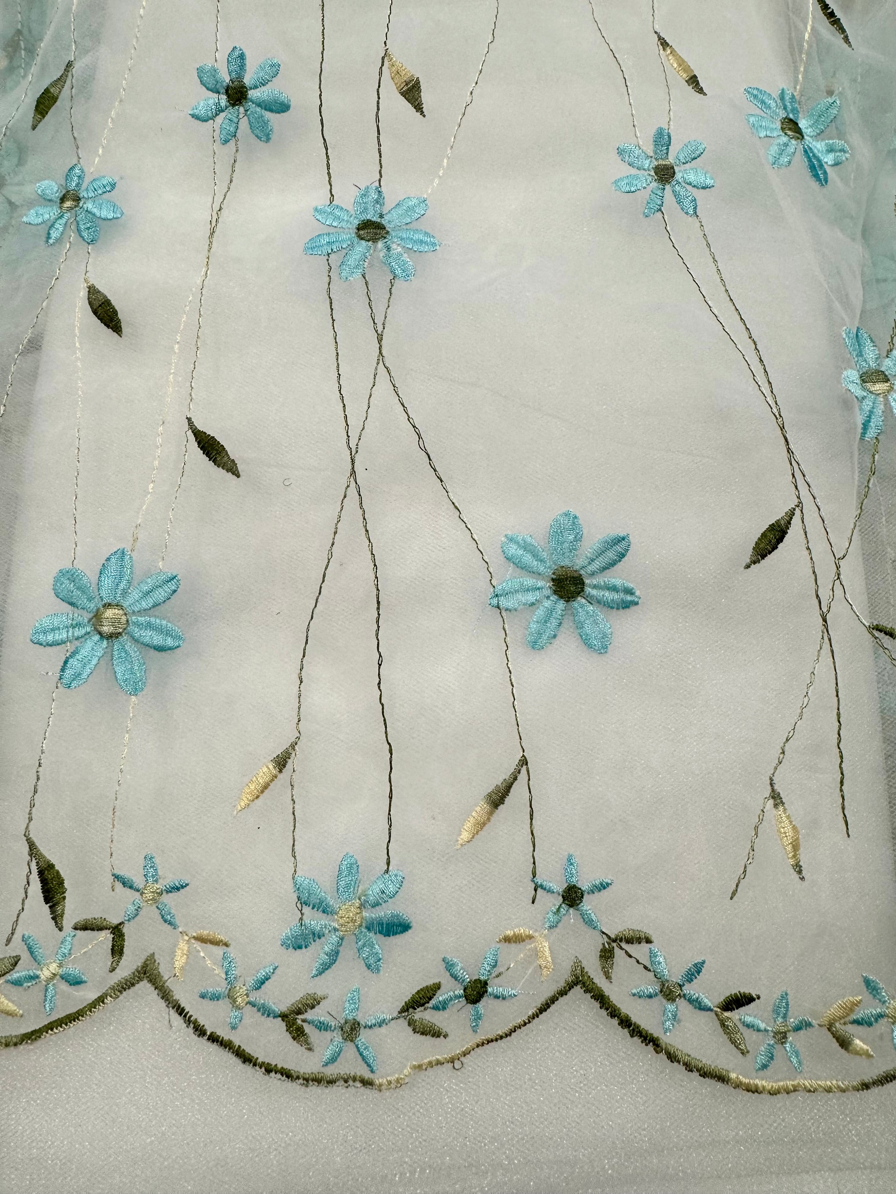 White sheer organza fabric with embroidered light blue and pale yellow flowers and green leaves, featuring a sage green scalloped embroidered border at the bottom.
