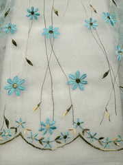 White sheer organza fabric with embroidered light blue and pale yellow flowers and green leaves, featuring a sage green scalloped embroidered border at the bottom.